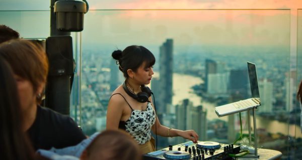 A DJ plays a rooftop gig in front of a city skyline at sunset. 