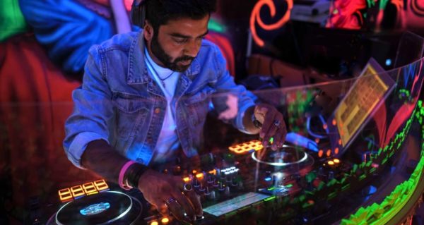 A DJ plays behind a glass screen in a vibrant, intimate nightclub setting. The wall decor appears to be black light activated. 