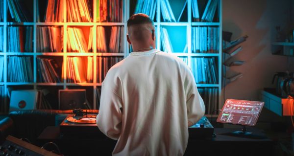 A DJ faces away from the camera, playing on turntables and a tablet, with Djay Pro and Spotify integration displayed. There's a wall filled with vinyl records along the back wall, and the lighting in the room is orange and blue. 