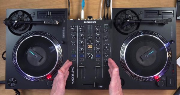 Top-down view of a DJ set-up on a wooden table in a well-lit room. There are two Reloop RP-7 turntables on each side of an Allen & Heath Xone:24C mixer. Two Phase devices are placed on each turntable. A person is gesturing with both hands to the mixer. 