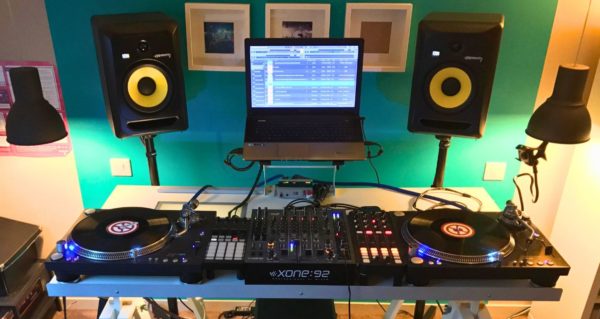A hybrid digital and vinyl DJ set-up in a warmly-lit studio. There's a laptop and two KRK speakers on stands against a bright green wall. 