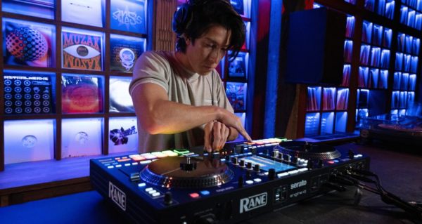 A young DJ mixing on a Rane System One standalone system in a room with dark blue lighting. Behind the DJ is a wall filled with vinyl records on display.