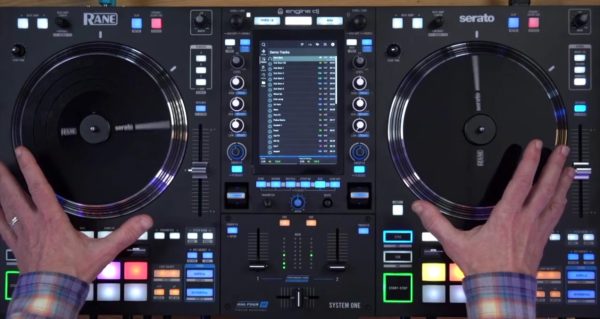 Overhead view of hands on the Rane System One's motorised platters, with the vertical touchscreen displaying a track list in Engine DJ between the two decks. RGB performance pads are illuminated at the bottom of each deck.