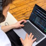 DJ Software: Who's Leading The Way In 2026? A person wearing headphones types on a laptop with Rekordbox DJ software, seated on a light-colored sofa against a wooden floor.