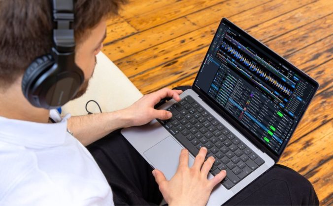 A person wearing headphones types on a laptop with Rekordbox DJ software, seated on a light-colored sofa against a wooden floor.