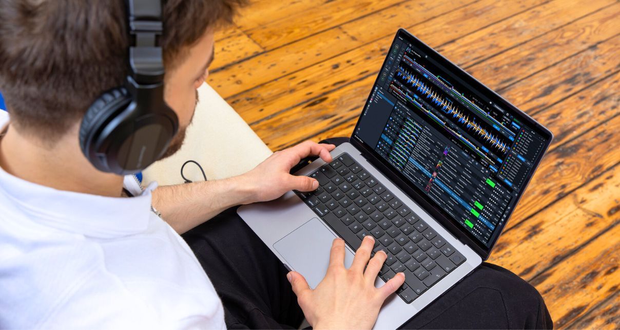 A person wearing headphones types on a laptop with Rekordbox DJ software, seated on a light-colored sofa against a wooden floor.