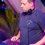 A DJ looks down at some CDJs, playing music in an outdoor tropical setting.