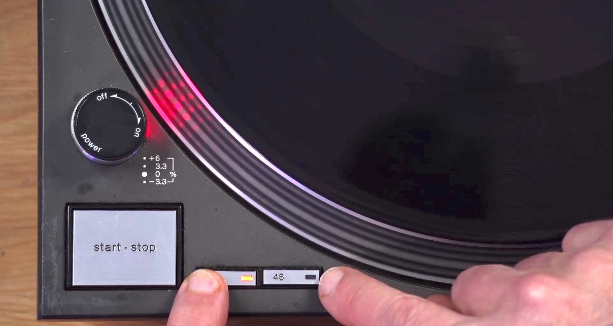 DJ's fingers pressing the 33 and 45 speed buttons on a Technics turntable with the strobe light showing red dots on the platter edge. 