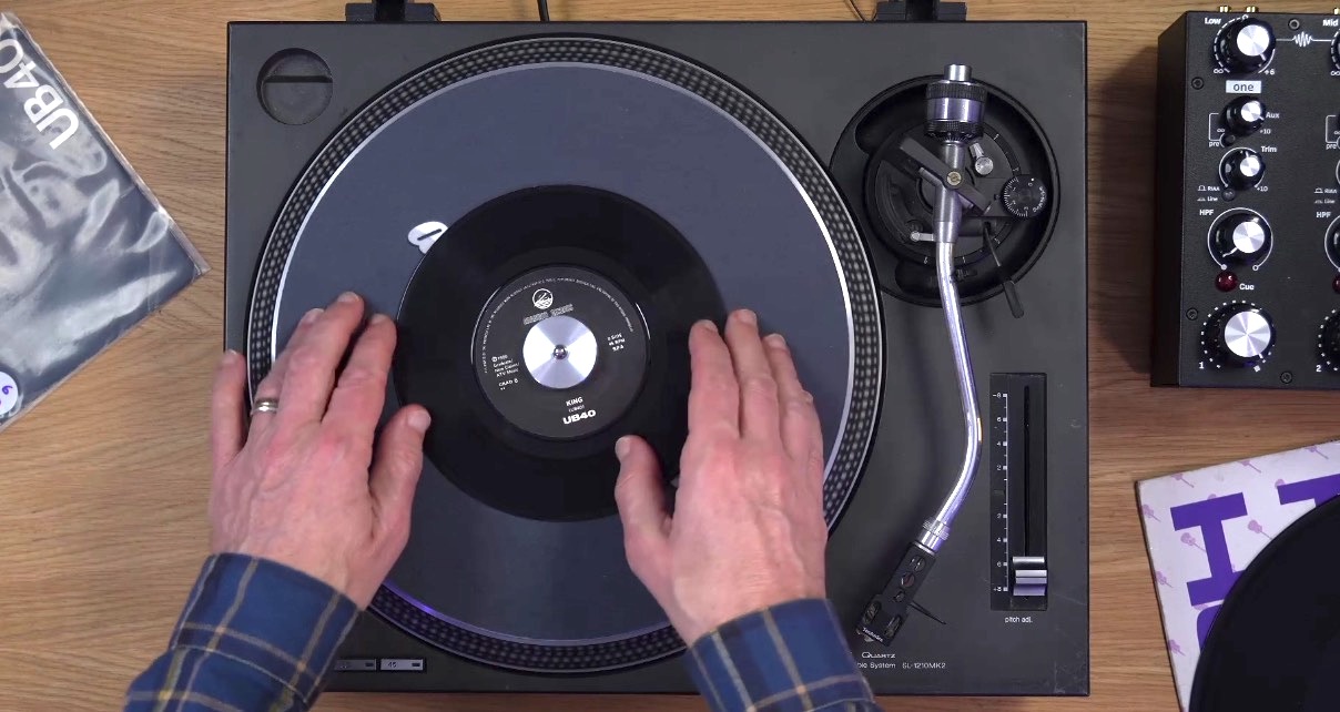 Overhead view of a Technics turntable with a 45 RPM record and centre adapter on the platter, with a DJ's hands positioned on either side of the deck. 