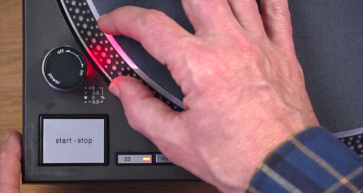 DJ's hand gesturing toward the strobe dots on the side of a spinning Technics turntable platter with red LED illumination.