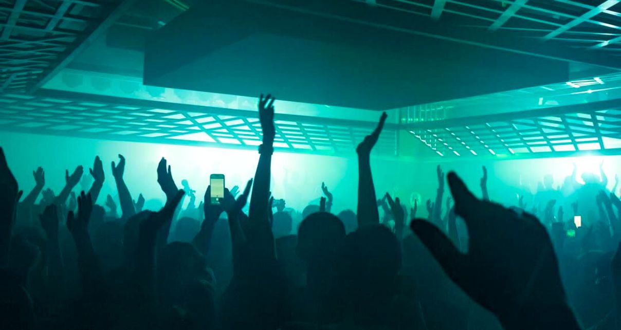 Crowd with hands raised dancing at Ministry of Sound's reopened Box room with new integrated lighting rig and dancefloor-level DJ booth. The photo is bathed in emerald green hues.