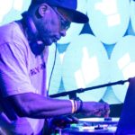 DJ Jazzy Jeff performing at a DJ booth wearing headphones and a baseball cap, adjusting controls on his equipment with a laptop visible, against a backdrop of turquoise and white thumbs-up icons on an LED screen.