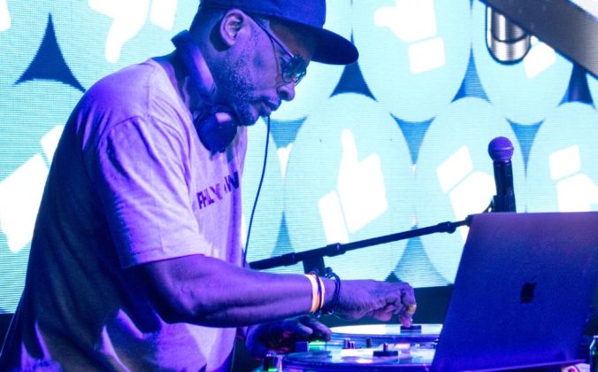 DJ Jazzy Jeff performing at a DJ booth wearing headphones and a baseball cap, adjusting controls on his equipment with a laptop visible, against a backdrop of turquoise and white thumbs-up icons on an LED screen.