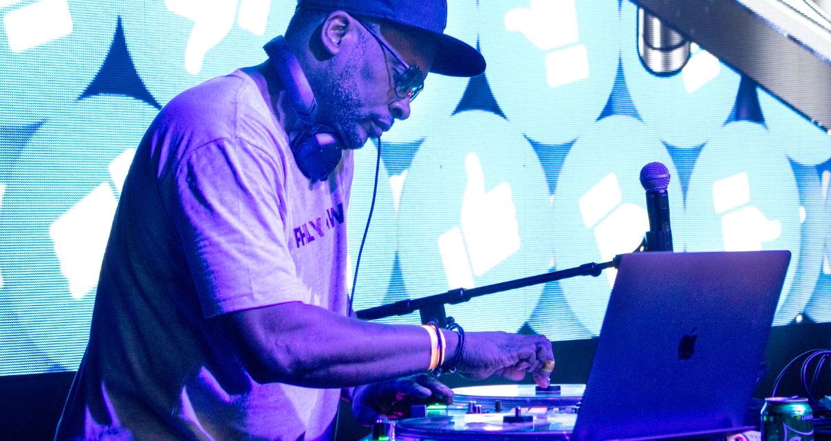 DJ Jazzy Jeff performing at a DJ booth wearing headphones and a baseball cap, adjusting controls on his equipment with a laptop visible, against a backdrop of turquoise and white thumbs-up icons on an LED screen.