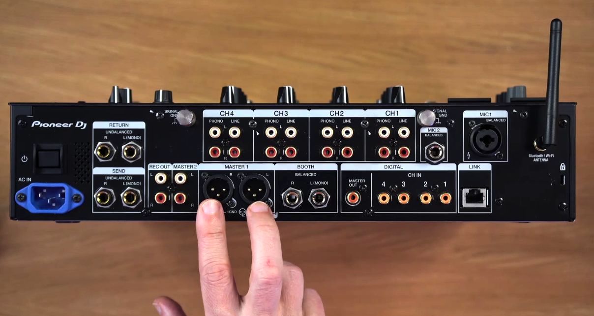 A person points at the Master outputs on a Pioneer DJ DJM-A9 mixer. The mixer is stood upright on a wooden desk in a well-lit room.
