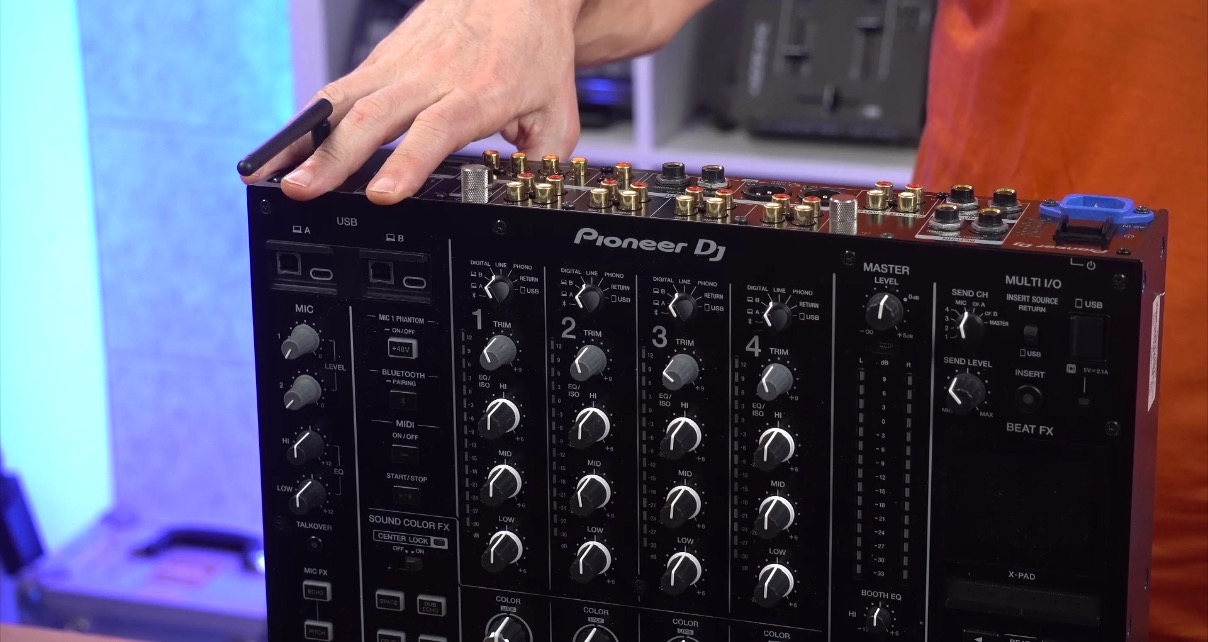 Hand pointing to USB-C and USB-B connections on the front panel of a Pioneer DJ DJM-A9 mixer with channel controls visible in the background.