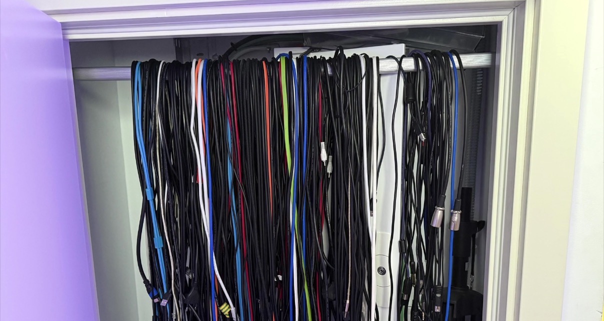 A large collection of DJ cables of various colours and types hanging loosely over a wardrobe rail inside a cupboard, with a purple-lit wall visible to the left.