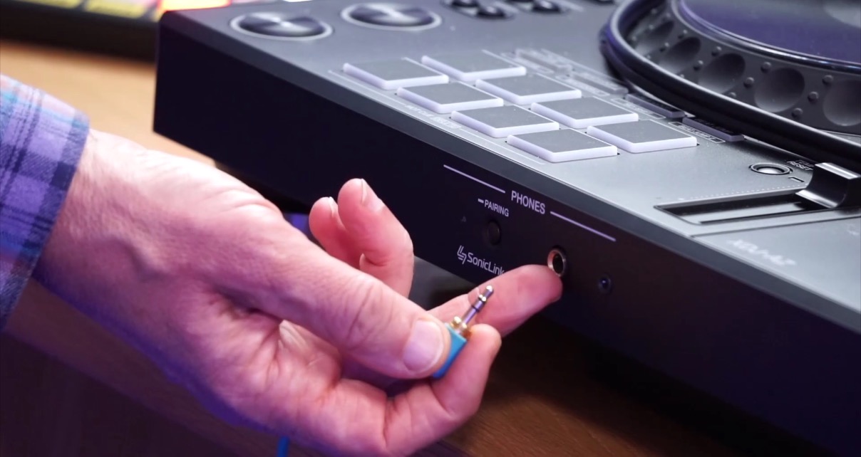 A hand holding a mini jack connector in front of the headphone section on a DJ controller, with both the quarter-inch and mini jack sockets visible alongside the "Phones" label.