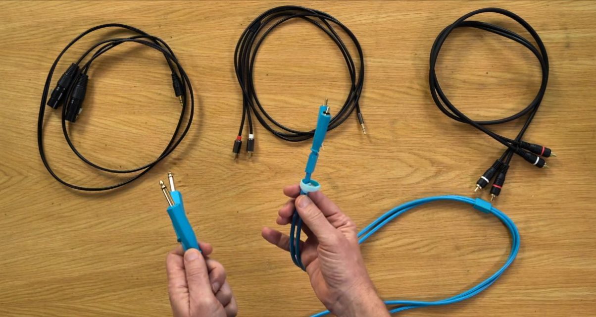 A pair of hands holding a blue cable with quarter-inch jack connectors, with three other coiled cables laid out on a wooden surface behind - including XLR, RCA, and mini jack to RCA cables.