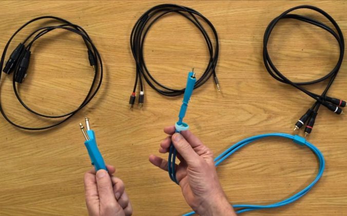 A pair of hands holding a blue cable with quarter-inch jack connectors, with three other coiled cables laid out on a wooden surface behind - including XLR, RCA, and mini jack to RCA cables.