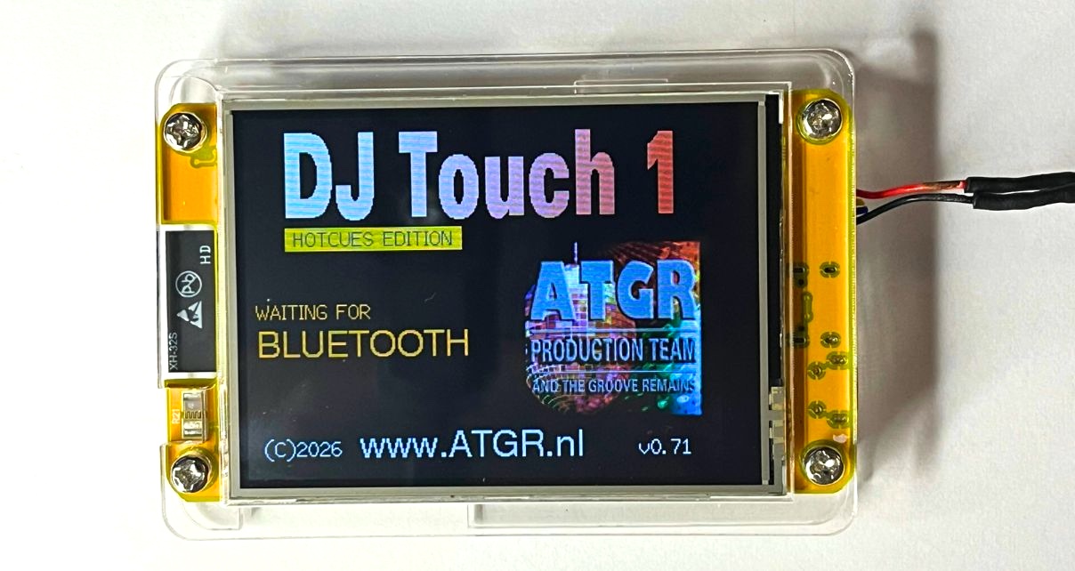 The original DJ Touch 1 in a clear plastic enclosure with yellow side brackets, showing its colour touchscreen displaying the boot screen with "DJ Touch 1 Hotcues Edition", "Waiting for Bluetooth", and the ATGR Production Team branding, with power wiring visible at the right side. 