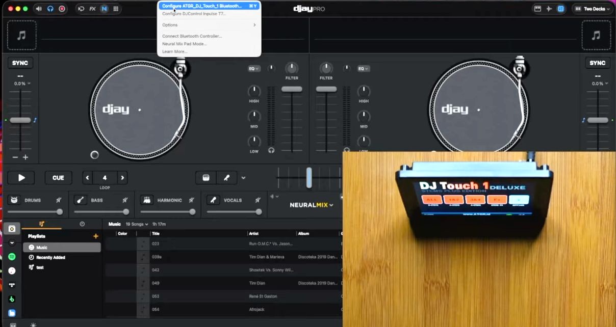 A screenshot of djay Pro on a Mac showing the DJ Touch 1 Deluxe being recognised as a Bluetooth controller, with a dropdown menu open displaying "Configure ATGR_DJ_Touch_1 Bluetooth..." as the top option, and the physical Deluxe unit visible in the bottom right corner of the screen showing its stems control interface. 