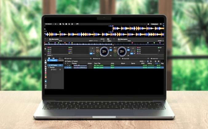 A silver laptop sits on a light wooden table displaying Rekordbox DJ software. The room is well lit with lots of windows and greenery in the background.