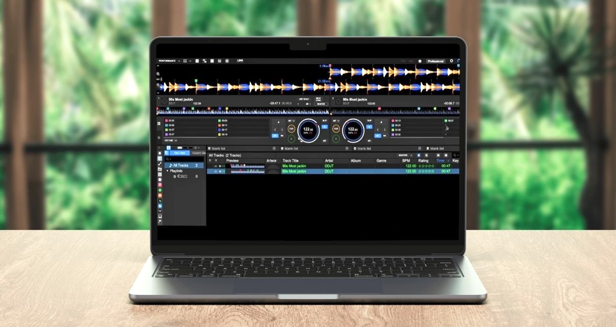 A silver laptop sits on a light wooden table displaying Rekordbox DJ software. The room is well lit with lots of windows and greenery in the background.