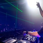 Ferry Corsten performing at a packed club, one arm raised to the crowd, green laser beams cutting through the darkness behind him.