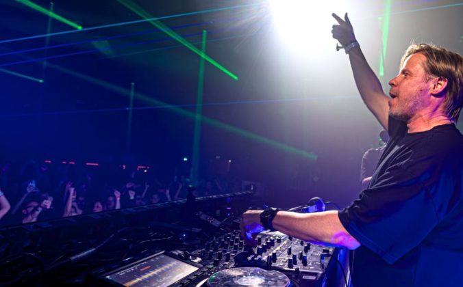 Ferry Corsten performing at a packed club, one arm raised to the crowd, green laser beams cutting through the darkness behind him.
