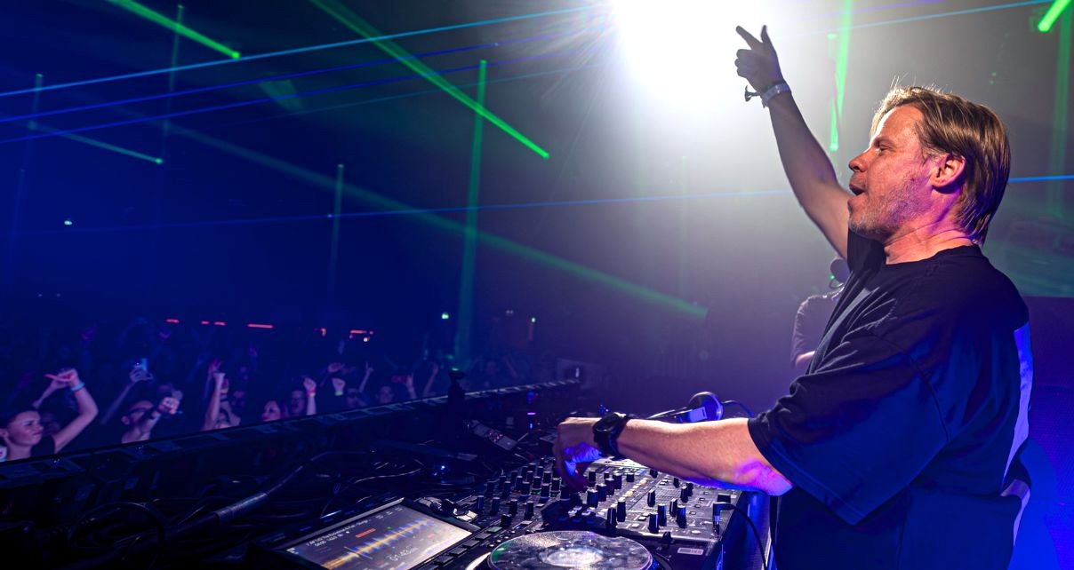 Ferry Corsten performing at a packed club, one arm raised to the crowd, green laser beams cutting through the darkness behind him.