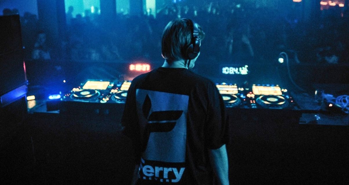 Ferry Corsten seen from behind at the DJ booth, wearing a branded Ferry Forever t-shirt, facing a dark crowded dancefloor lit by blue light, with CDJs and a mixer spread across the decks in front of him. 